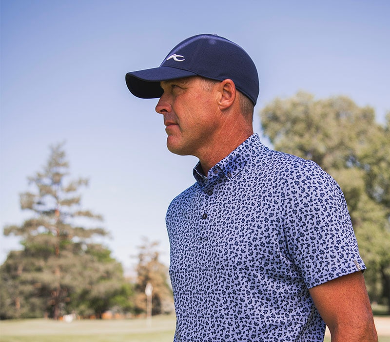 Golfer wearing KJUS printed polo and a navy cap standing outside with trees around him.